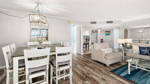Un vaste espace de vie comprenant une table à manger, des chaises blanches et un canapé. Un lustre est suspendu au plafond et un tableau orne le mur.