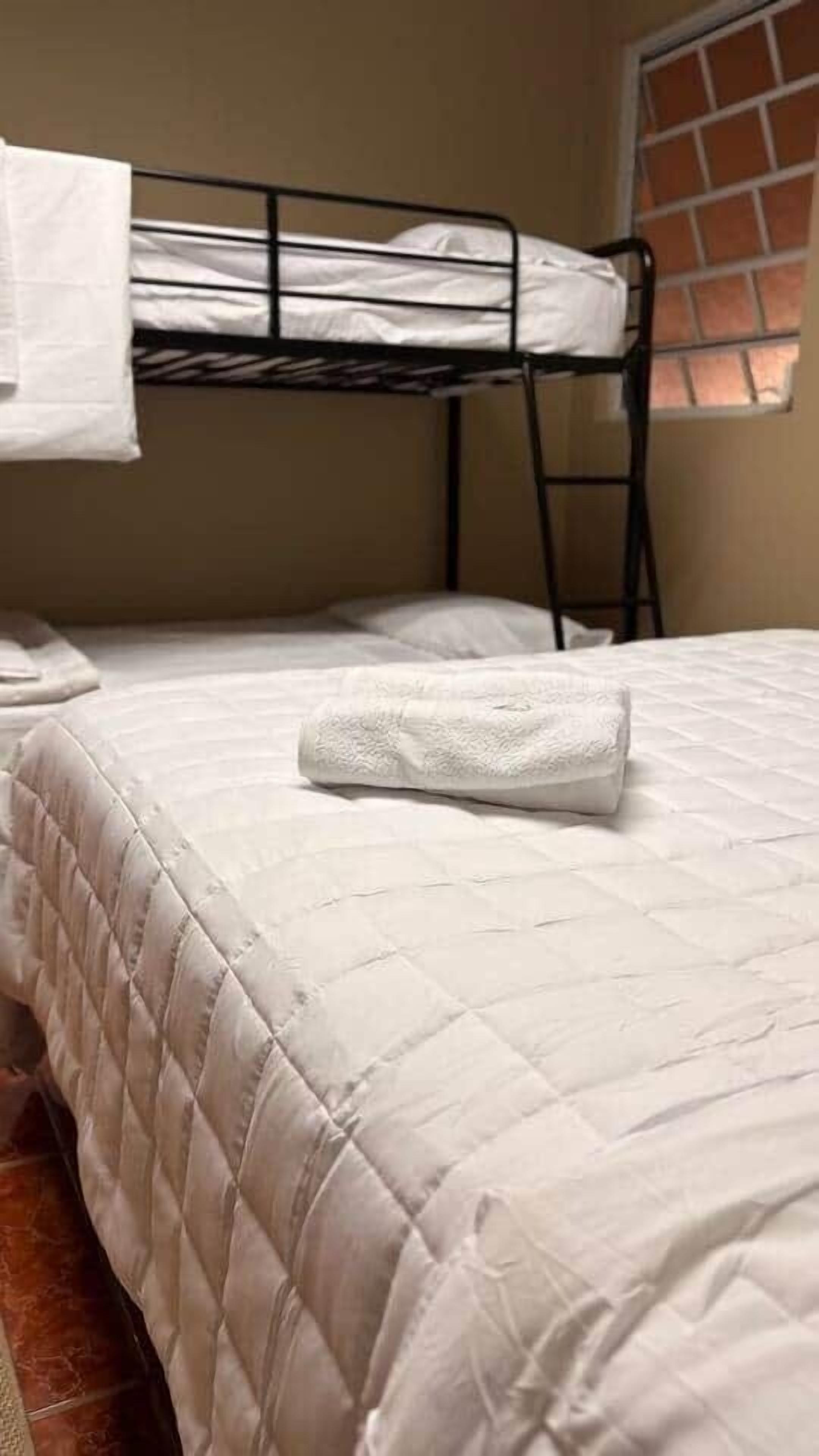 Una cama individual con edredón blanco acolchado, una litera metálica encima y una ventana con vistas al exterior.