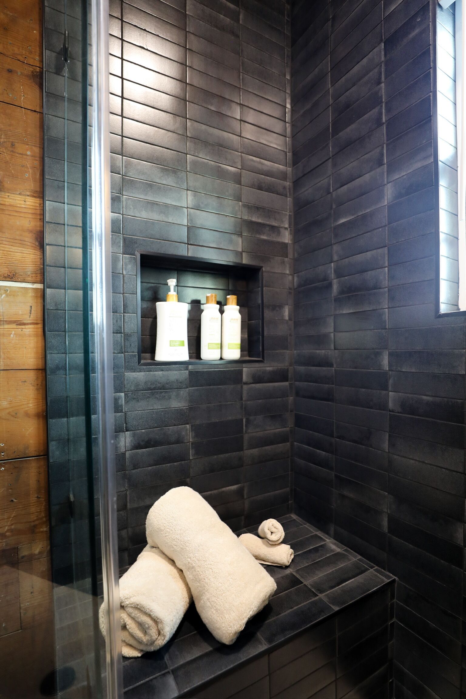 A modern bathroom with dark tiled walls, a glass shower enclosure, and a shelf with toiletries.