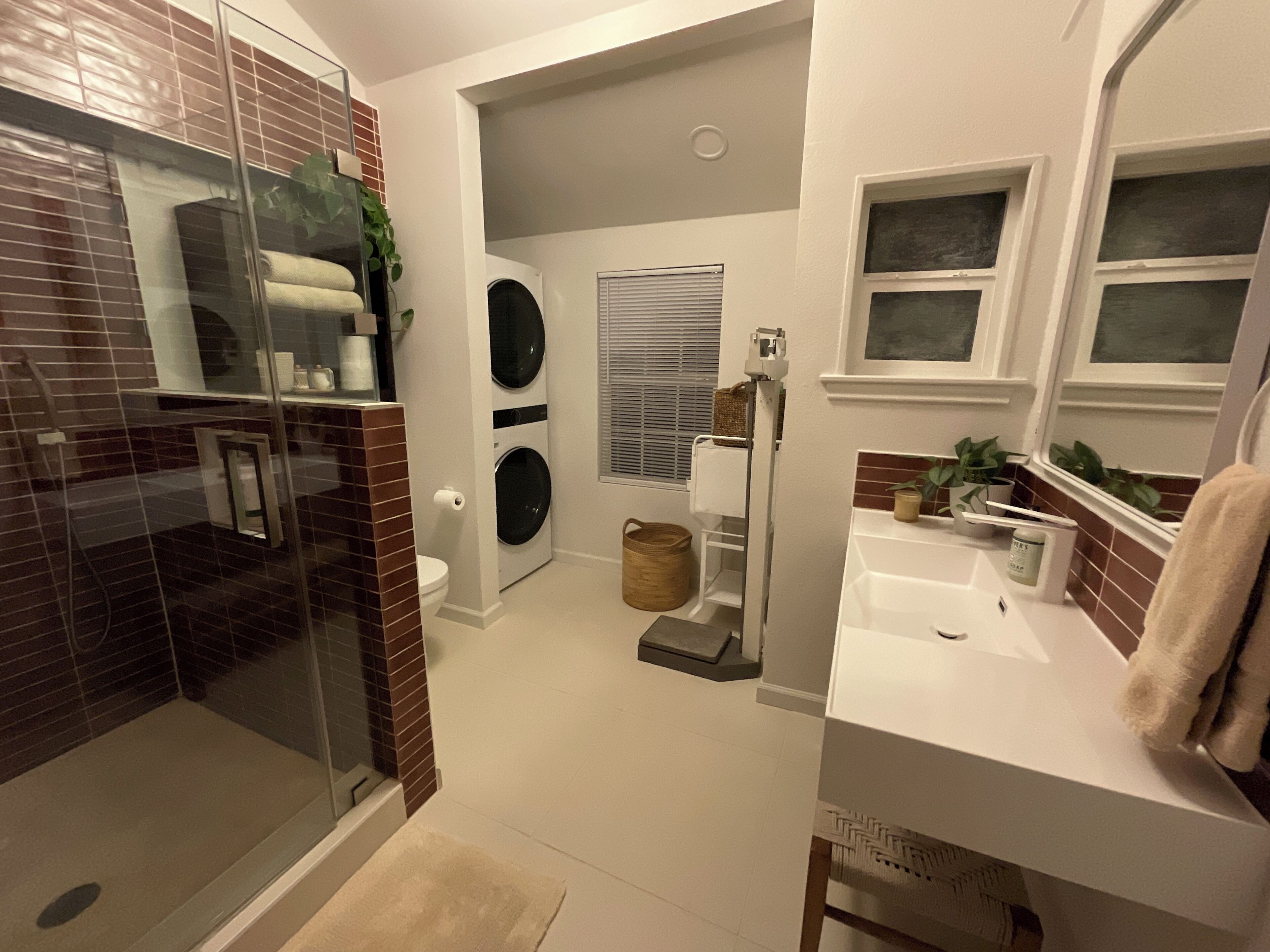 A compact bathroom with a glass shower enclosure, a washing machine, and a sink with a mirror.