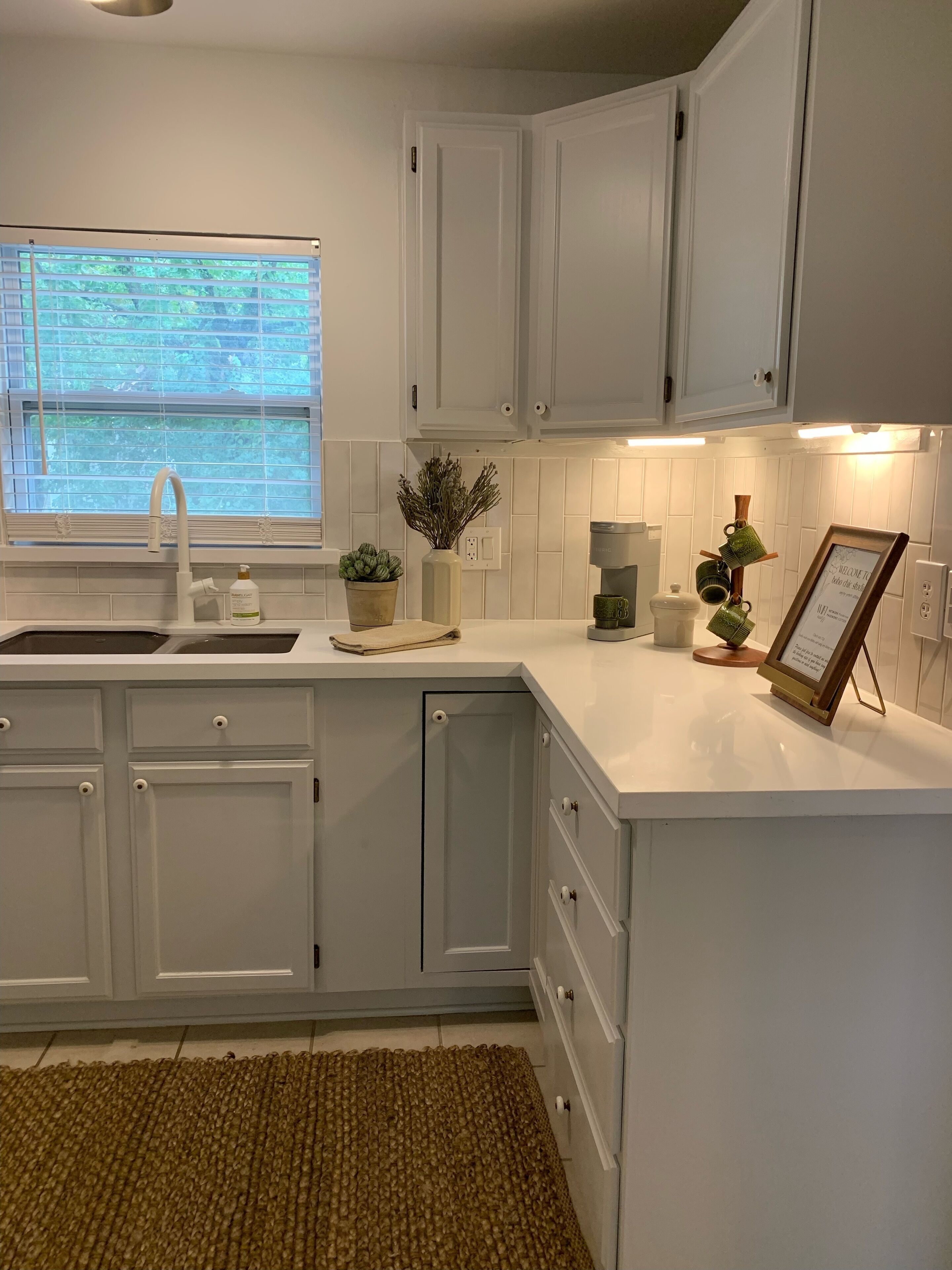 A kitchen with white cabinets, a sink, a window with blinds, and a small potted plant.