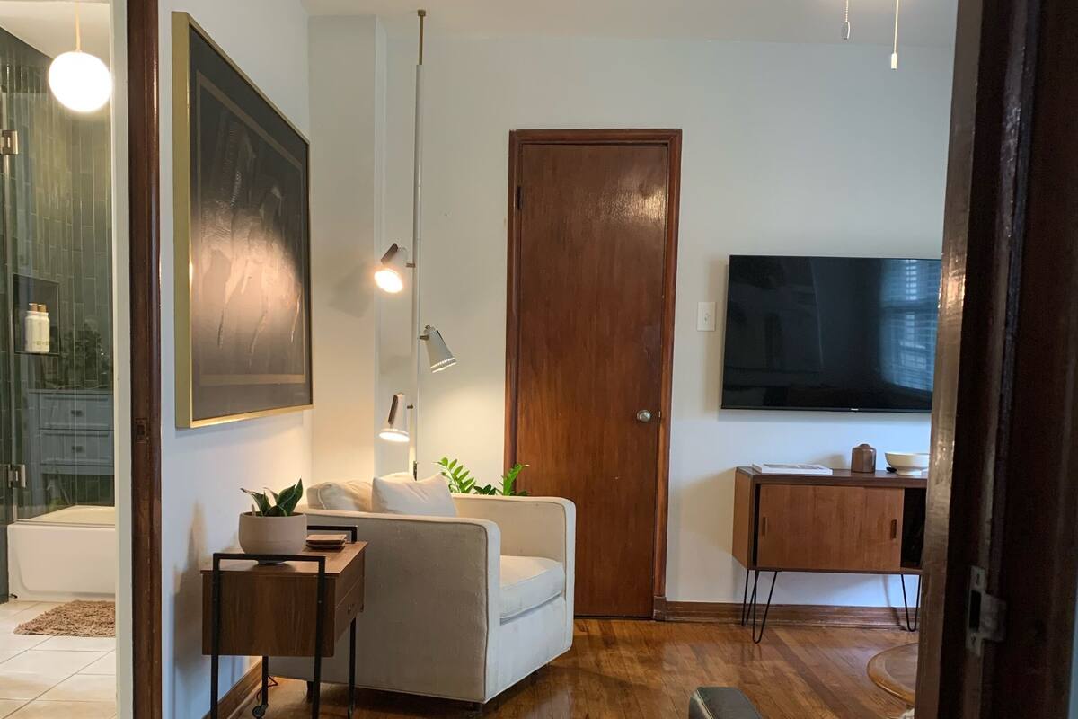 Un salon moderne avec un plancher en bois, un canapé blanc, une petite table sur laquelle est posée une plante, un ventilateur de plafond et un téléviseur fixé au mur.