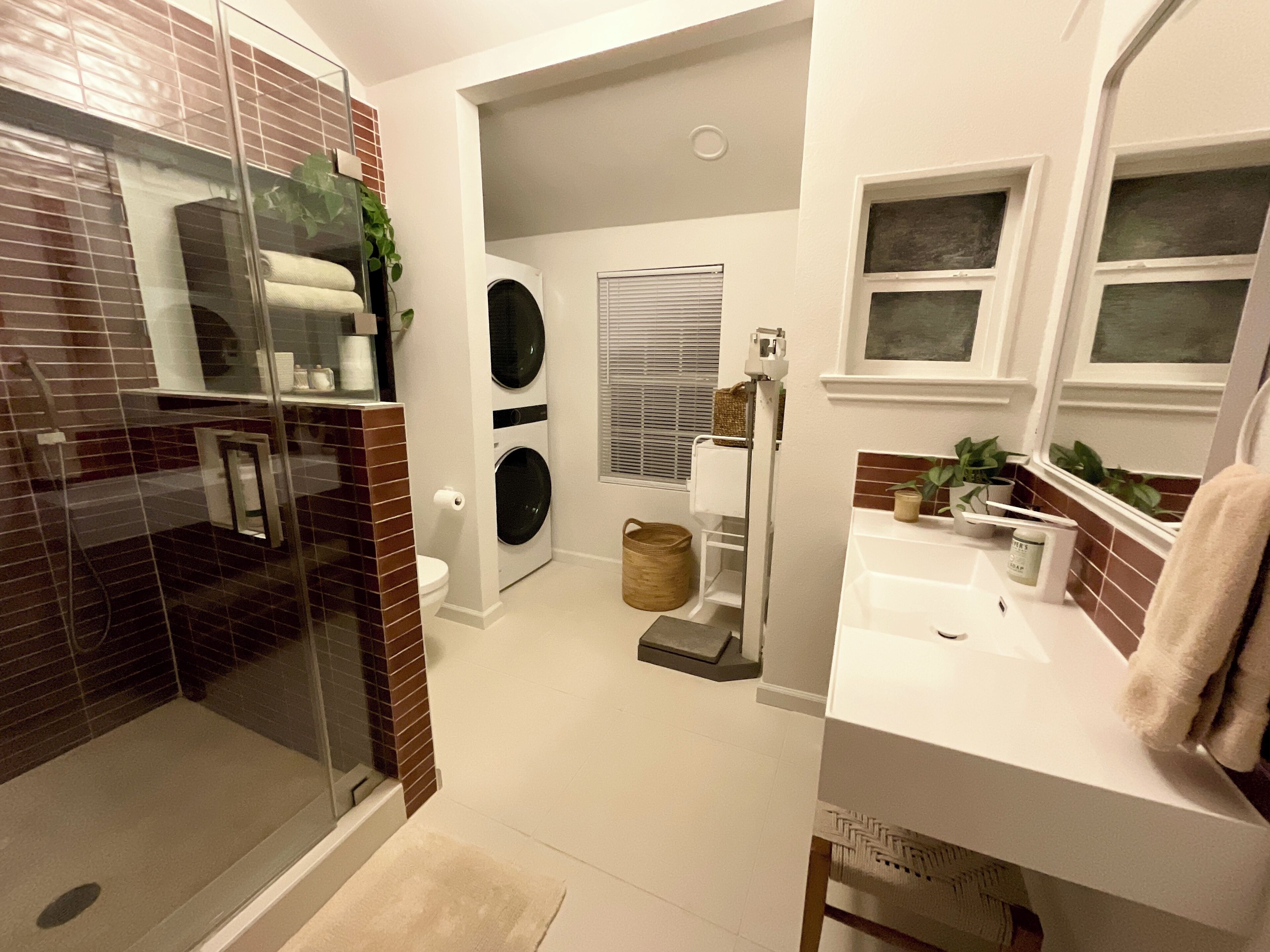 A modern bathroom with a glass shower enclosure, a built-in washer and dryer, a white sink, and a mirror with shelves.