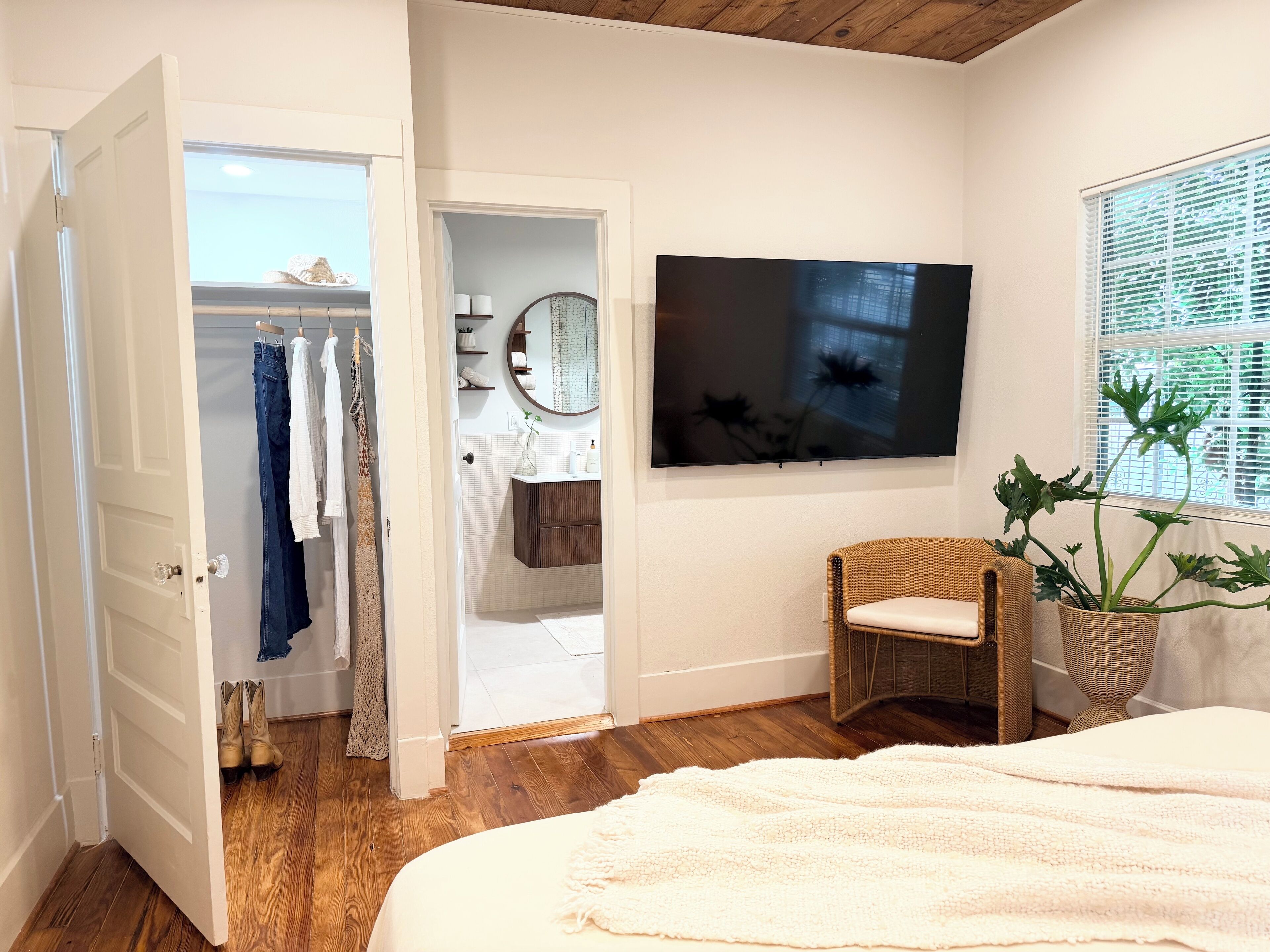 A bedroom with a bed, a TV, a wicker chair, a potted plant, and a closet with hanging clothes.