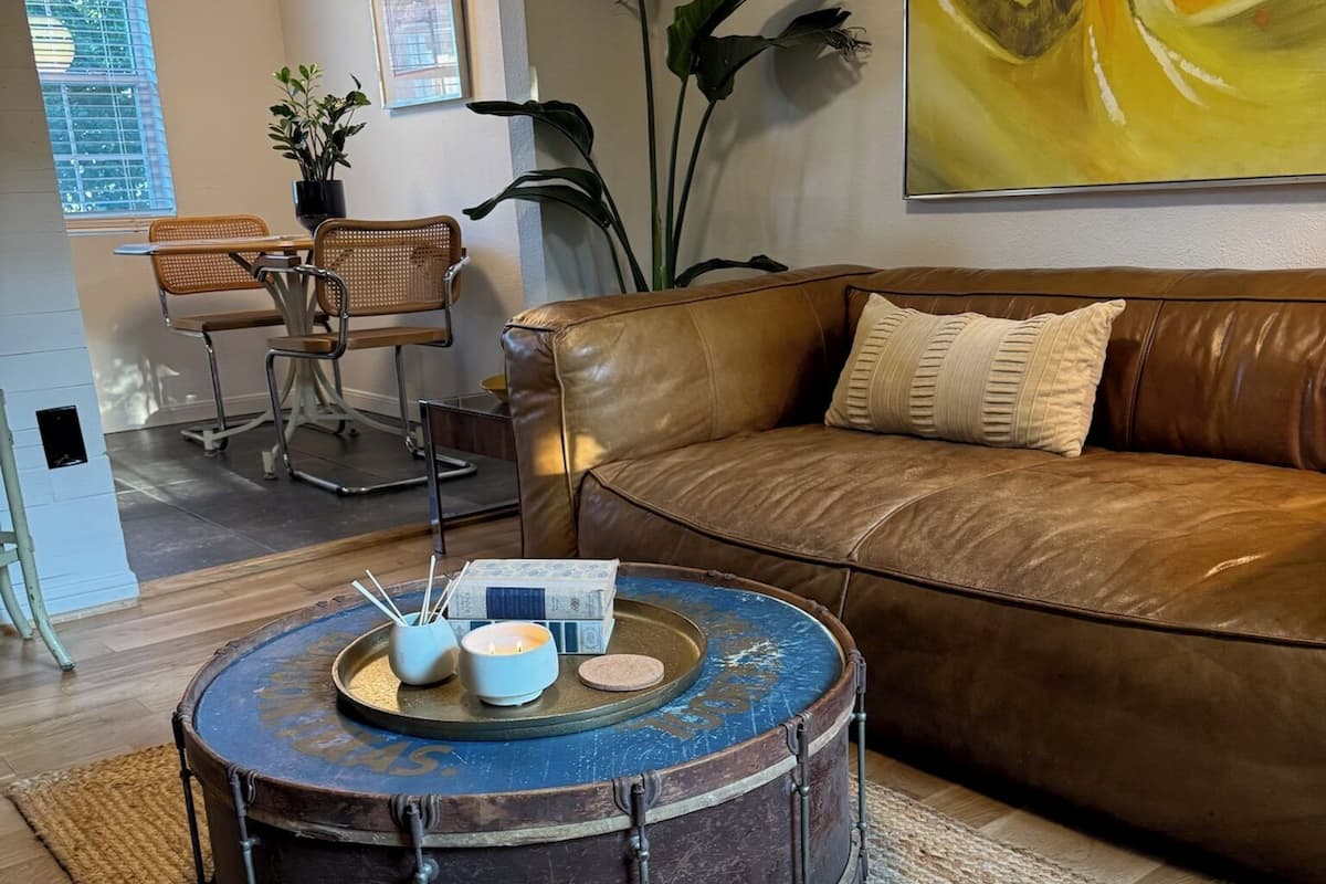 Un salon avec un canapé en cuir marron, une table ronde en forme de tambour décorée de bougies, un tapis tissé et un coin repas avec des chaises et une table en osier.