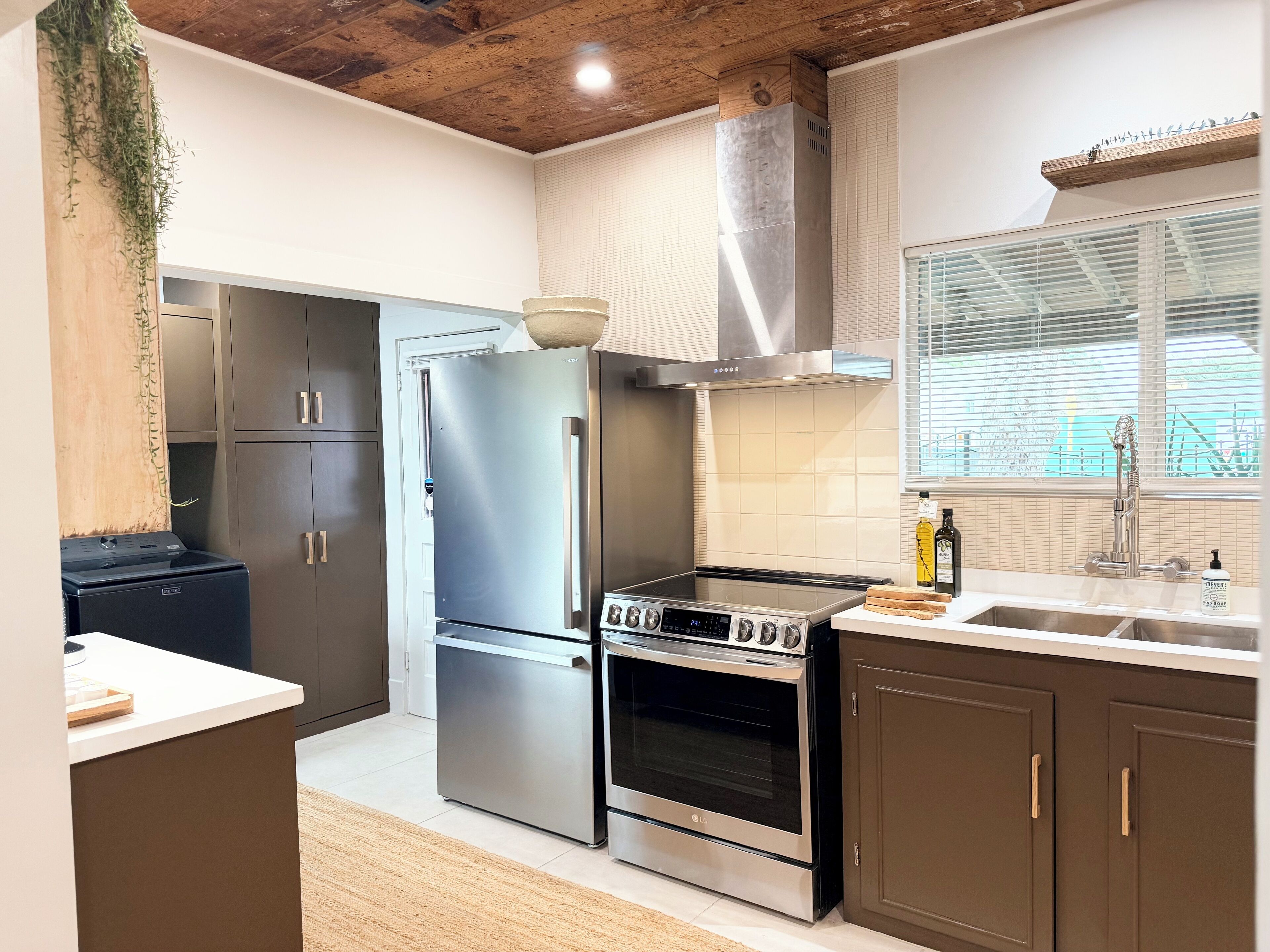 A modern kitchen with stainless steel appliances, wooden cabinets, and a large window with blinds.