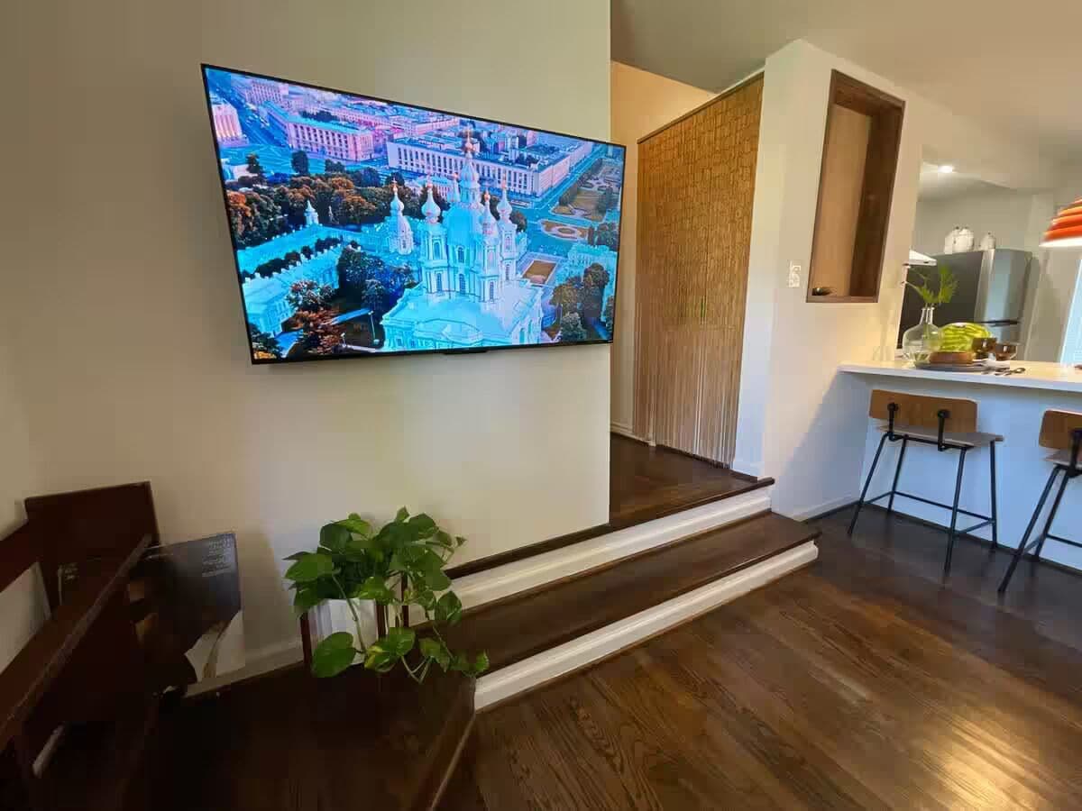 Un salon moderne avec un grand téléviseur à écran plat fixé au mur, un escalier en bois, une petite plante et un coin cuisine équipé d’un réfrigérateur et de tabourets de bar.