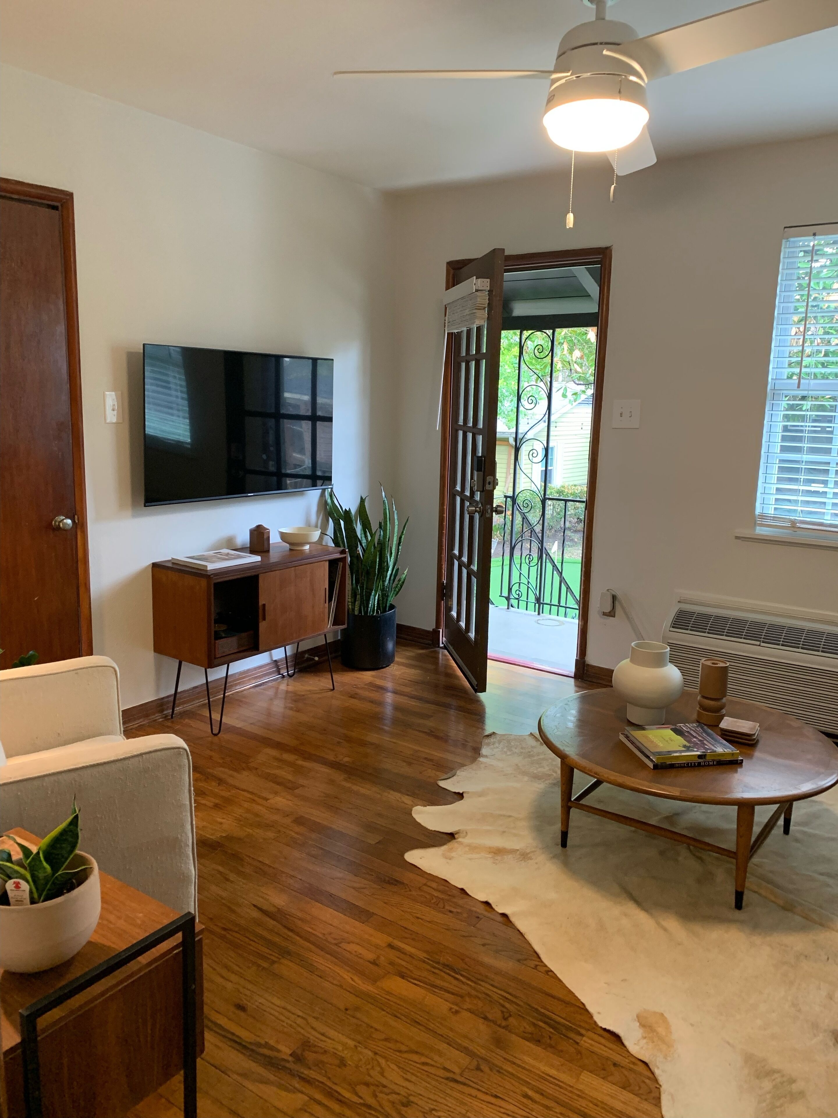 A modern living room with a flat-screen TV, wooden furniture, a ceiling fan, and a large window with blinds.