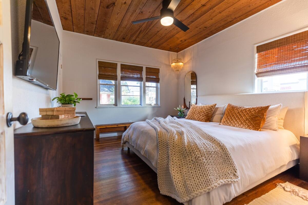 Une chambre avec un plafond en bois, un lit avec une literie blanche, une table de chevet, un miroir et une fenêtre avec des stores.