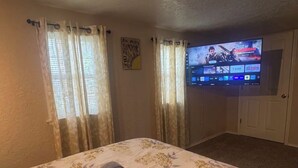 A hotel room with a bed, curtains, a television displaying a movie, and a motivational quote on the wall. - Cozy, FUN home! Game room with pool table and arcade game 5-7 min from Fort Sill (Lawton)