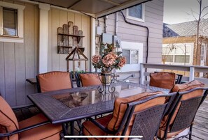 Een terras buiten met een tafel met glazen blad, gestoffeerde stoelen, een decoratieve houten plank en een bloevas.