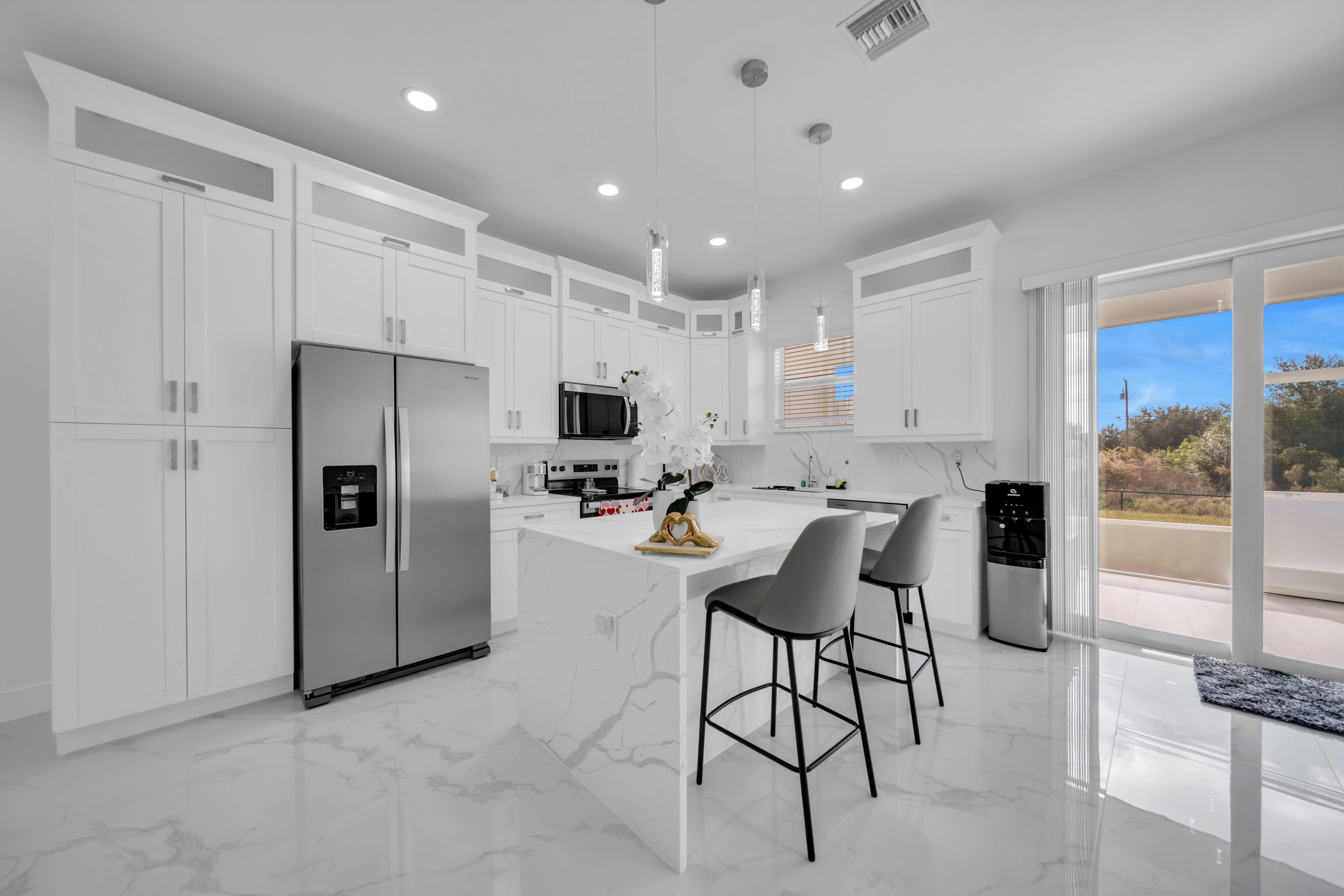 Une cuisine moderne avec des meubles blancs, un îlot central doté de tabourets de bar, des appareils en acier inoxydable et une vue sur l’extérieur grâce à de grandes fenêtres.