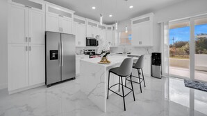 A modern kitchen with white cabinetry, a central island with bar stools, stainless steel appliances, and a view of the outdoors through large windows. - Brand New 3BR Home Near Fort Myers Airport – Private & Modern (Lehigh Acres)
