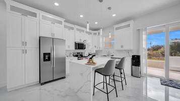 Une cuisine moderne avec des meubles blancs, un îlot central doté de tabourets de bar, des appareils en acier inoxydable et une vue sur l’extérieur grâce à de grandes fenêtres.
