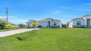 A residential area with three houses, a paved driveway, and a well-maintained lawn. - Brand New 3BR Home Near Fort Myers Airport – Private & Modern (Lehigh Acres)