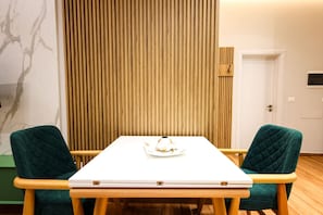 Una habitación con suelo de madera, una mesa blanca con una computadora portátil, dos sillas verdes, un panel de pared de madera y una puerta.