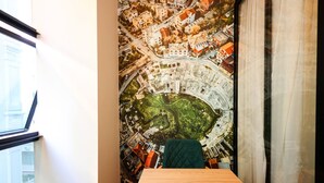 Ett rum med ett stort fönster, utsikt över en väggmålning som föreställer en stadsbild, ett träbord och en grön stol.