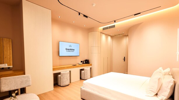 A modern hotel room with a bed, a desk, a chair, a TV displaying 'Grandstay Collection', and a wardrobe. - Zins Luxury Room Vollga (Durrës)