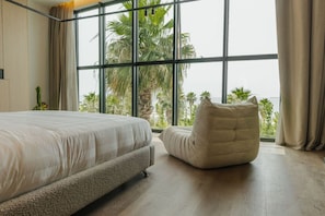 A bedroom with a large bed, a cushioned chair, and a view of palm trees through large windows. - Zins Luxury Apartment 01 (Durrës)