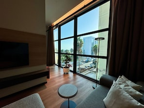 Uma sala de estar moderna com uma janela grande, uma televisão de tela plana, um sofá, uma mesa redonda pequena e uma planta em vaso.