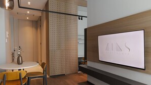 Een modern interieur met een tv waarop het logo van 'AINS APARTMENTS' staat, een ronde eettafel met stoelen, en een houten scheidingswand met een geperforeerd ontwerp.