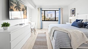 A modern bedroom with a large bed, a desk, and a view of the city through a window. - Gorgeous Cocoa Beach Condo: Balcony, Heated Pool, BBQ Grills & Hot Tub ☀️ (Cocoa Beach)