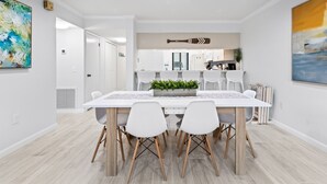 A modern dining area with a white table and chairs, a kitchen in the background, and a large abstract painting on the wall. - Gorgeous Cocoa Beach Condo: Balcony, Heated Pool, BBQ Grills & Hot Tub ☀️ (Cocoa Beach)