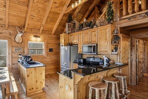 A wooden cabin interior with a kitchen featuring a refrigerator, microwave, and stove, a wooden dining table, and a bar area with stools. - Mtn Views, FIrepit, Hot Tub, Porch Swing, Pet-Friendly, Sleeps 12 (Sevierville)
