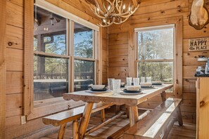 A wooden cabin interior with a dining table set for four, large windows, and a chandelier. - Mtn Views, FIrepit, Hot Tub, Porch Swing, Pet-Friendly, Sleeps 12 (Sevierville)