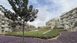 A residential complex with modern architecture, a central lawn, and purple flowering plants. - Elegant 2-bedroom Serviced with Aprt Pool view in Sodic Villette (Cairo Governorate)