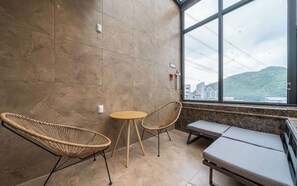 Une pièce dotée d’une grande fenêtre, de deux fauteuils en osier, d’une petite table ronde et d’un banc.