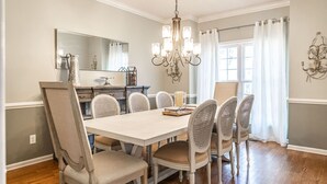 Uma sala de jantar com uma mesa grande, seis cadeiras, um candelabro, um espelho e uma janela com cortinas.