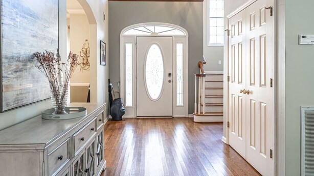 Um corredor com piso de madeira, uma porta branca com painéis de vidro, uma escada com corrimão branco e um vaso com flores secas sobre um console.