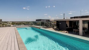 En pool på taket med ett trädäck, ett avskärmat sällskapsrum med glasväggar och utsikt över stadsbilden.