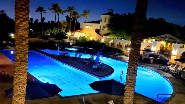 Une piscine entourée de palmiers, avec des chaises longues et des parasols, et un bâtiment dont les fenêtres sont illuminées.
