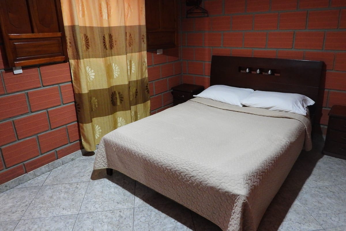 A bedroom with a bed, a nightstand, a wooden wardrobe, and a brick wall.