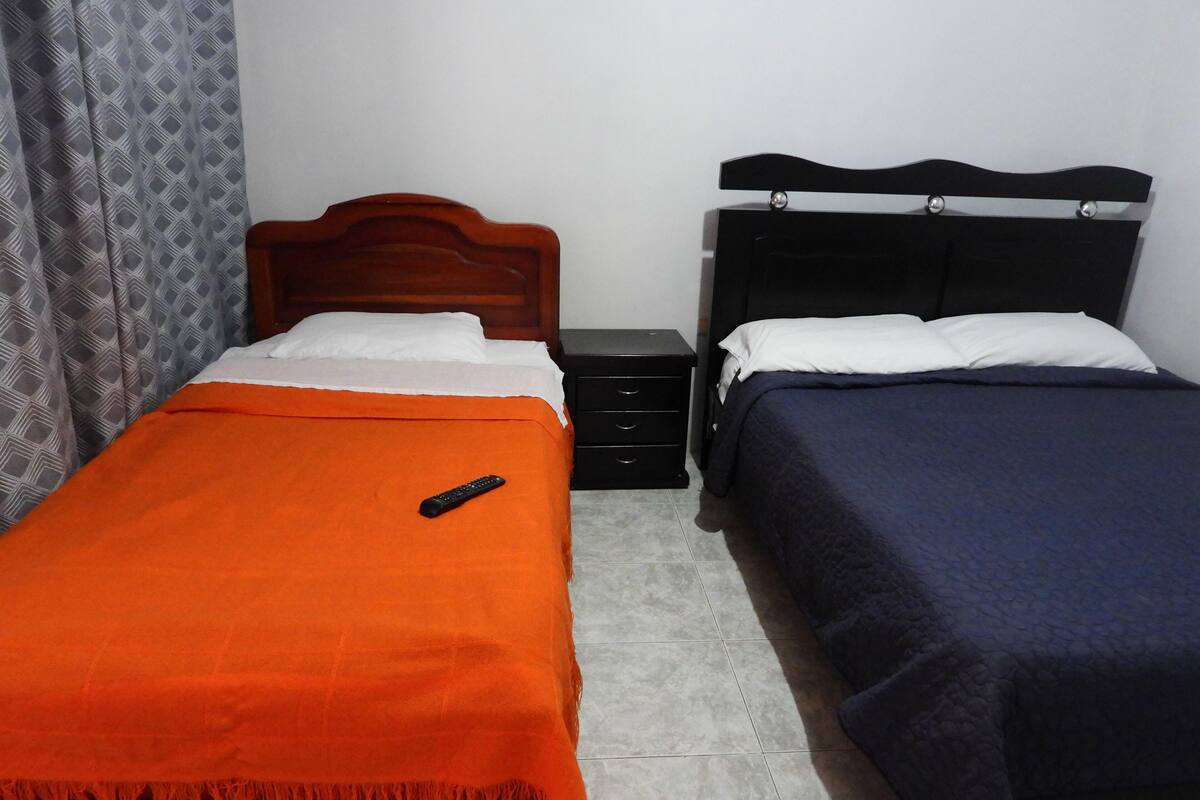 A room with two single beds, each with a different colored bedspread, a nightstand with a drawer, and a remote control on the left bed.
