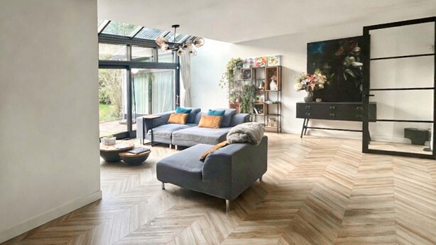 Una sala moderna con piso de patrón espiga, un sofá seccional gris, una mesa de centro y una estantería con plantas.