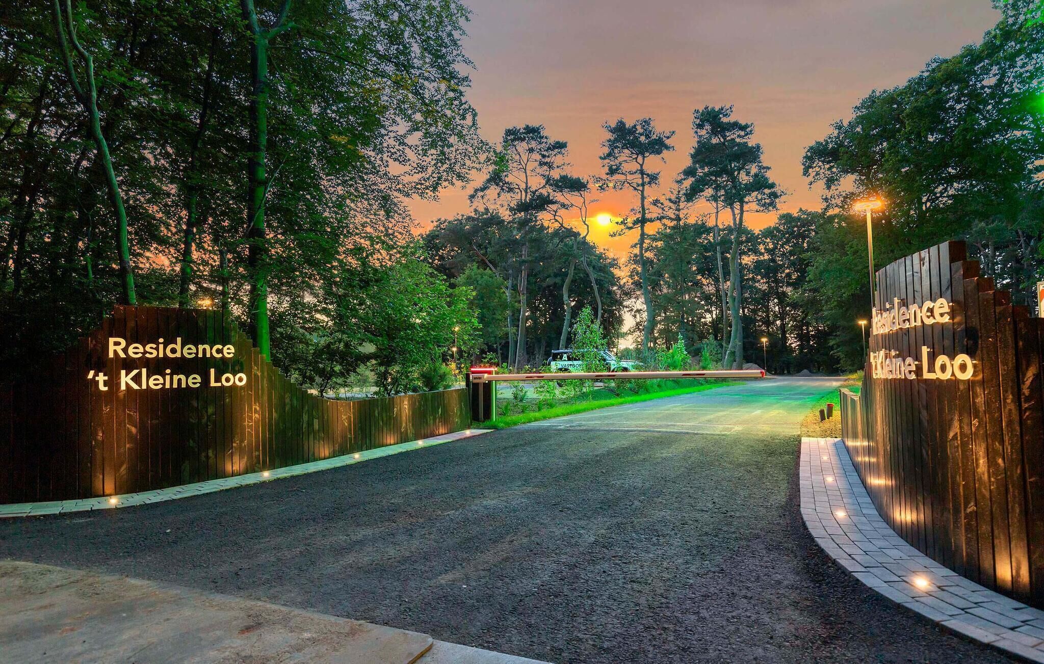 A gated entrance with 'Residence Kleine Loo' and 'Residence: Laine Loo' signs, surrounded by trees and a sunset in the background.