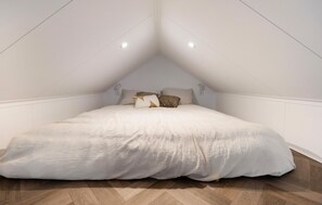 Een slaapkamer met een schuine kap, een bed met witte dekbedovertrek, twee nachtlampjes en een houten vloer.