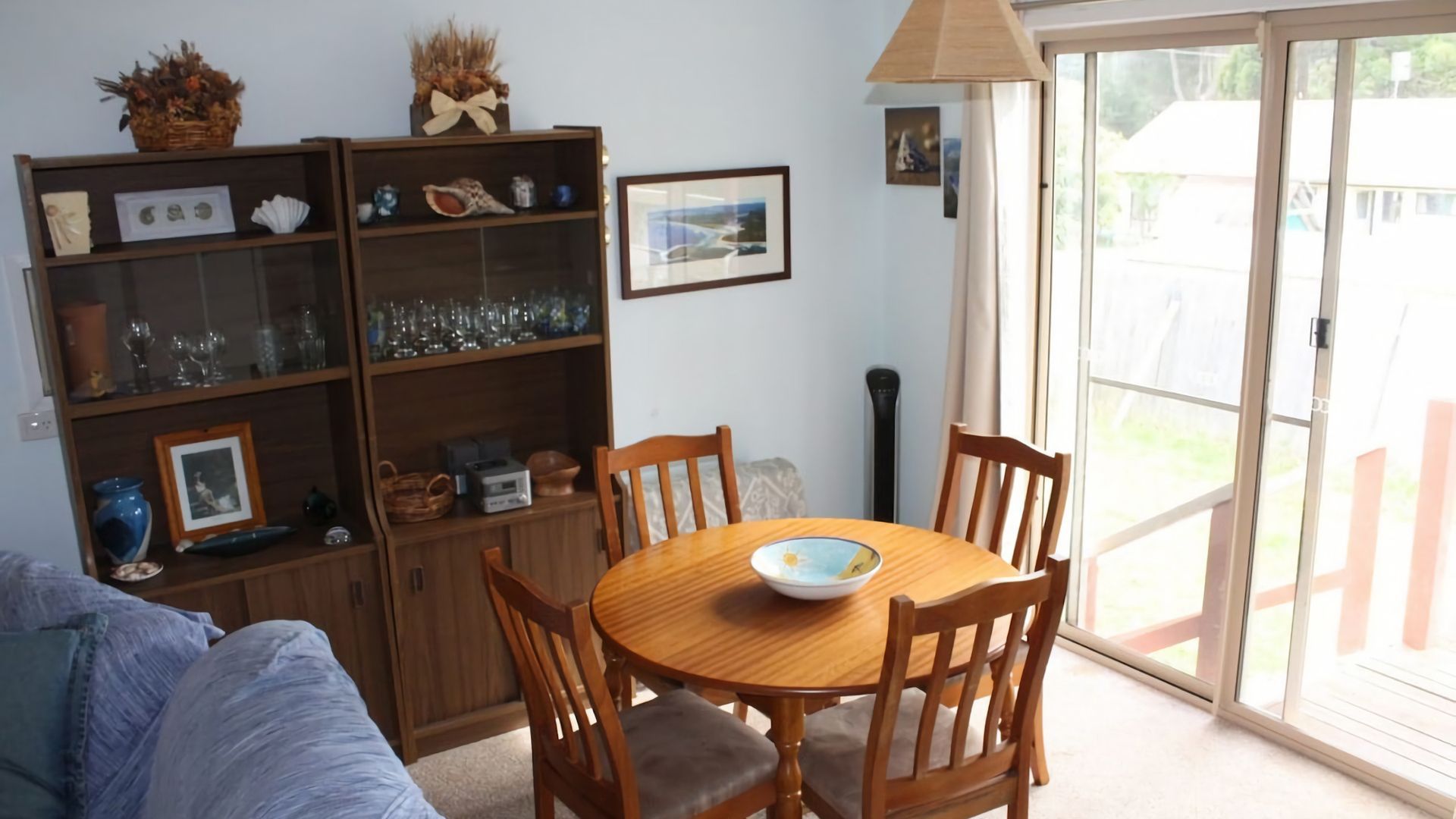 Un coin repas avec une table et des chaises en bois, une bibliothèque contenant divers objets et une porte-fenêtre coulissante donnant sur un espace extérieur.