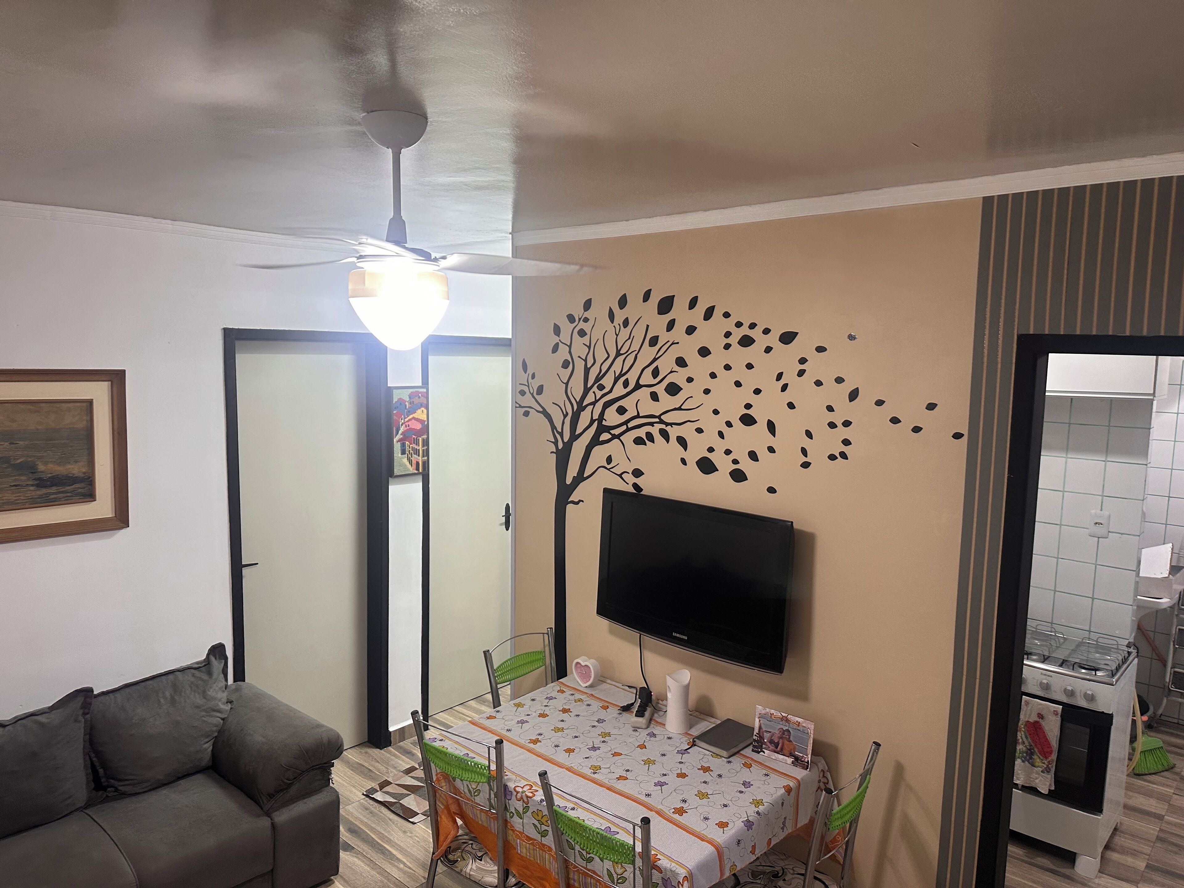 A living room with a sofa, a dining table with chairs, a flat-screen TV, a ceiling fan, and a wall-mounted decoration.