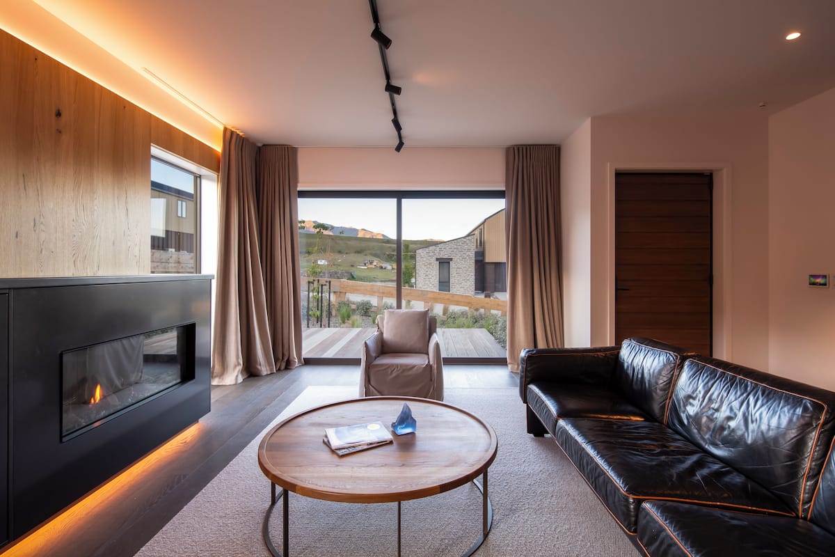 A modern living room with a fireplace, a leather sofa, a round coffee table, and a view of a hillside through large windows.