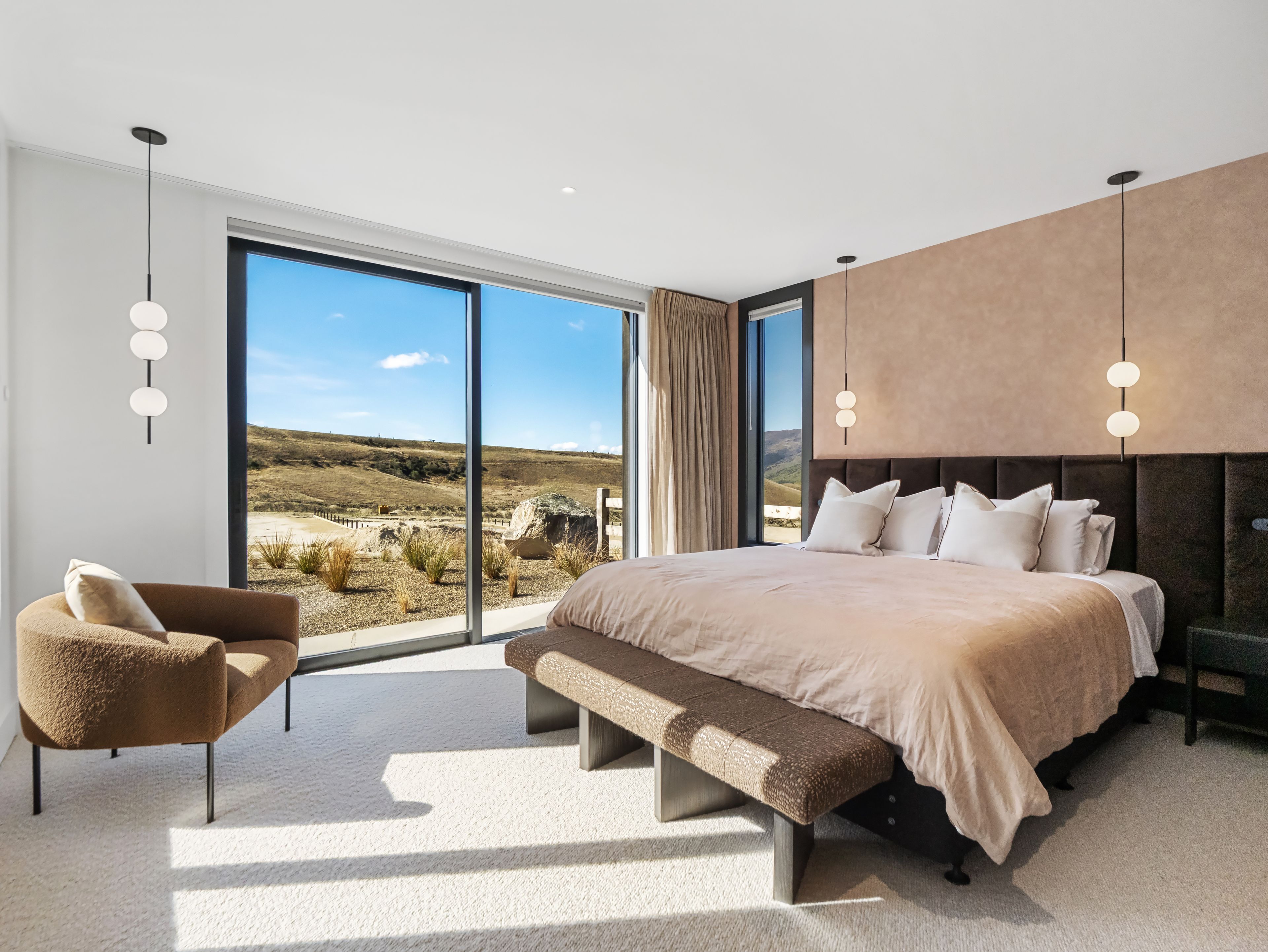 A modern bedroom with a large bed, a bench, a chair, and a view of a desert landscape through large windows.