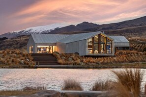 A modern house with large windows, a metal roof, and a view of a lake and mountains.