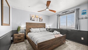 Une chambre à coucher avec un lit en bois, un ventilateur de plafond, une fenêtre avec des stores et des œuvres d’art accrochées au mur.