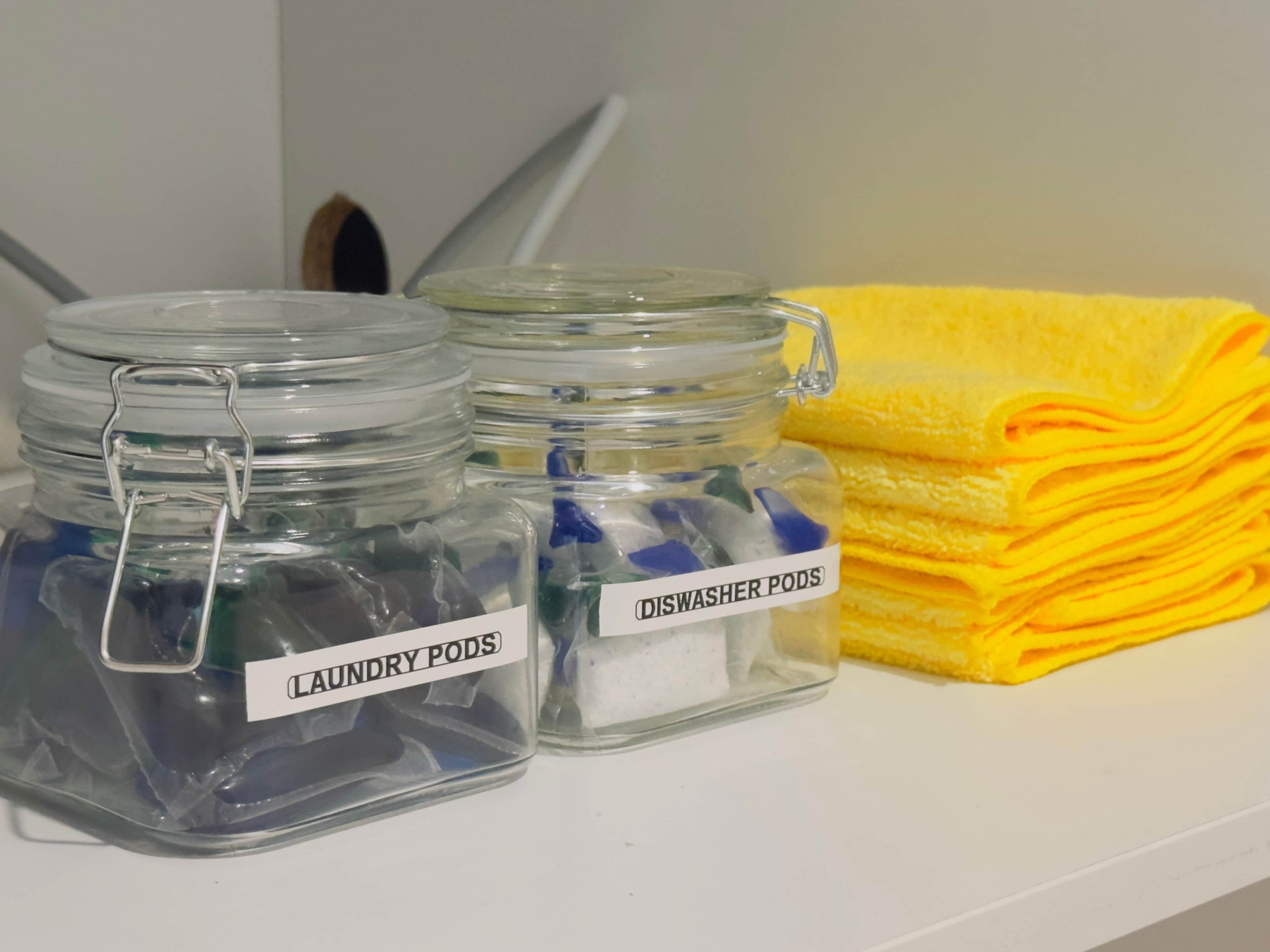 Three glass jars labeled 'laundry pods' and 'dishwasher pods' on a white surface with a yellow towel.