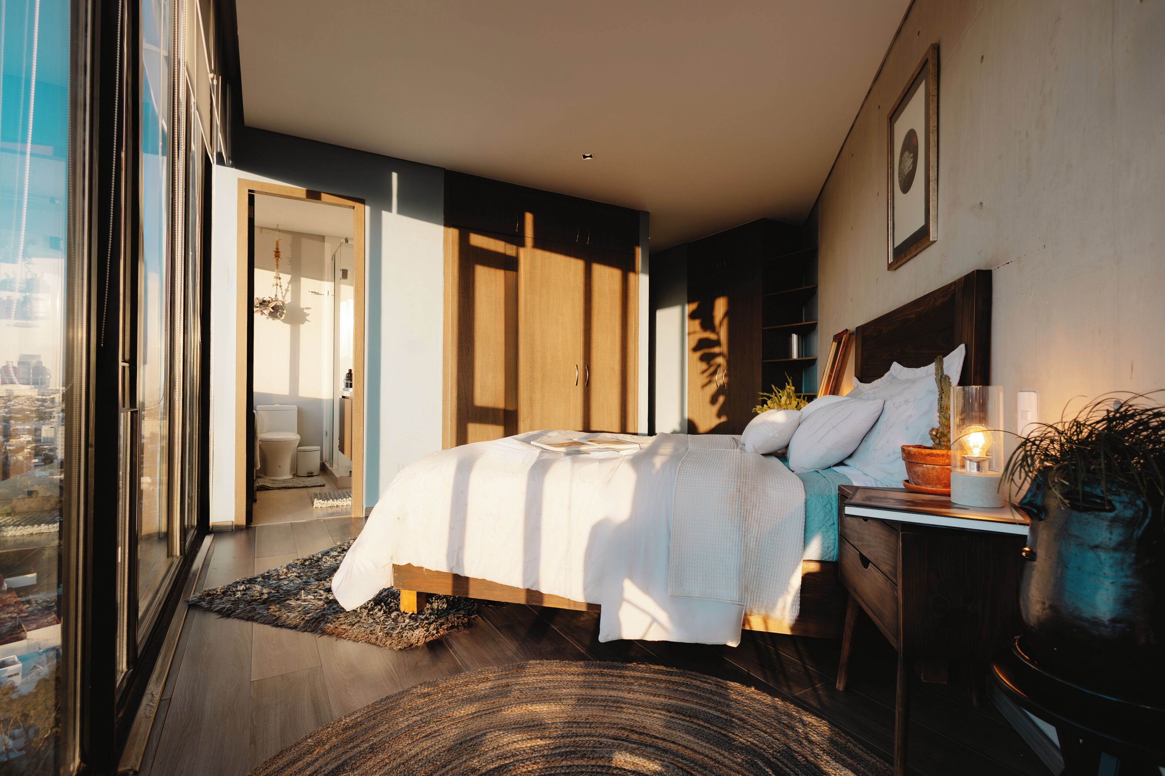 Een moderne slaapkamer met een groot bed, een nachtkastje met een lamp, en uitzicht op de stad door middel van raam-tot-plafond ramen.