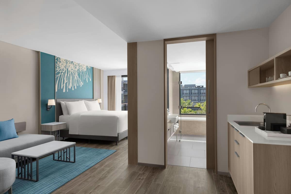 Een moderne hotelkamer met een bed, een bank, een tafeltje, een kitchenette en een balkon met uitzicht over de stad.