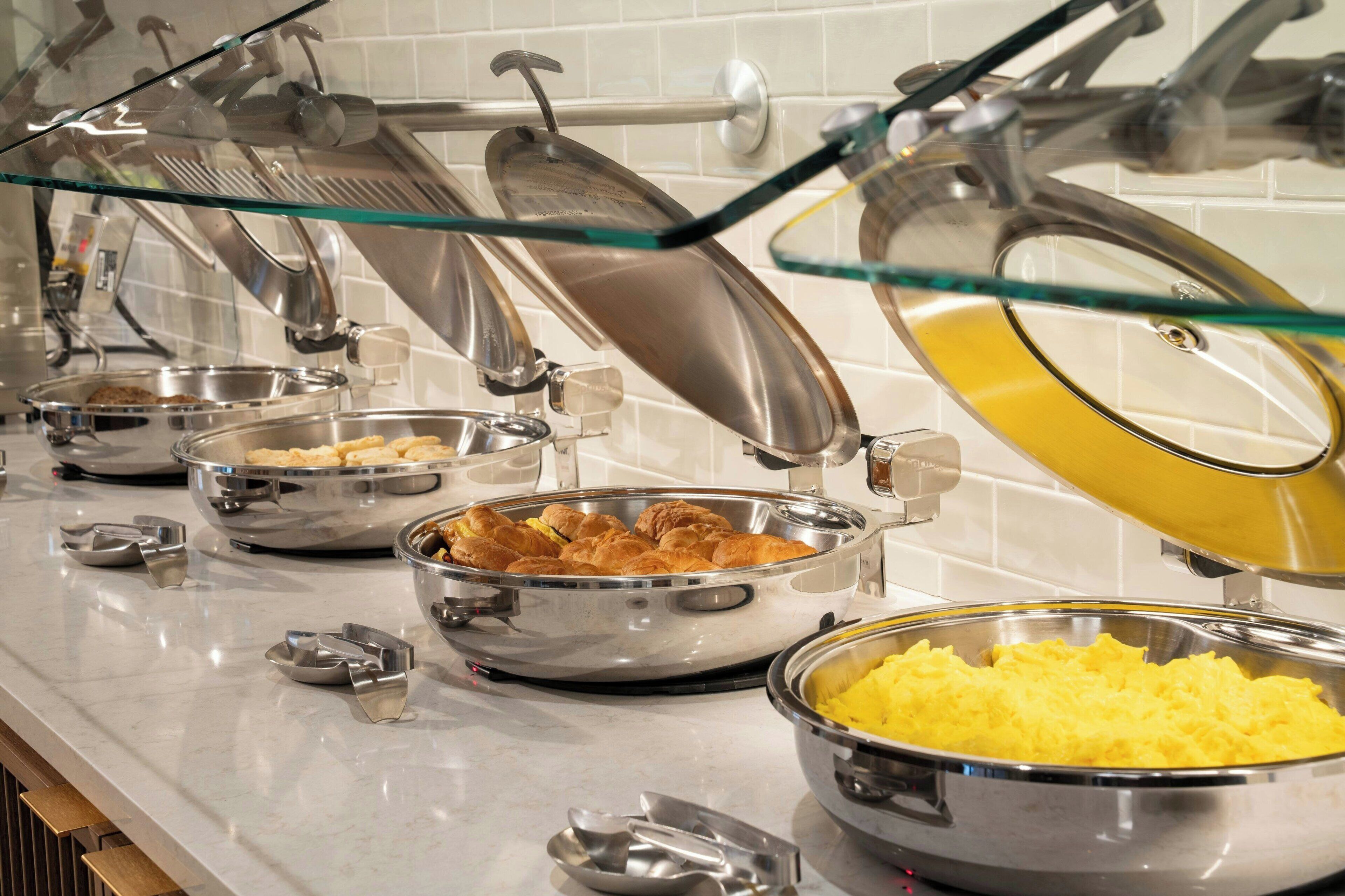 Ein reichhaltiges Buffet mit verschiedenen Speisen in Metallschüsseln, darunter offenbar Rühreier und Gebäck.